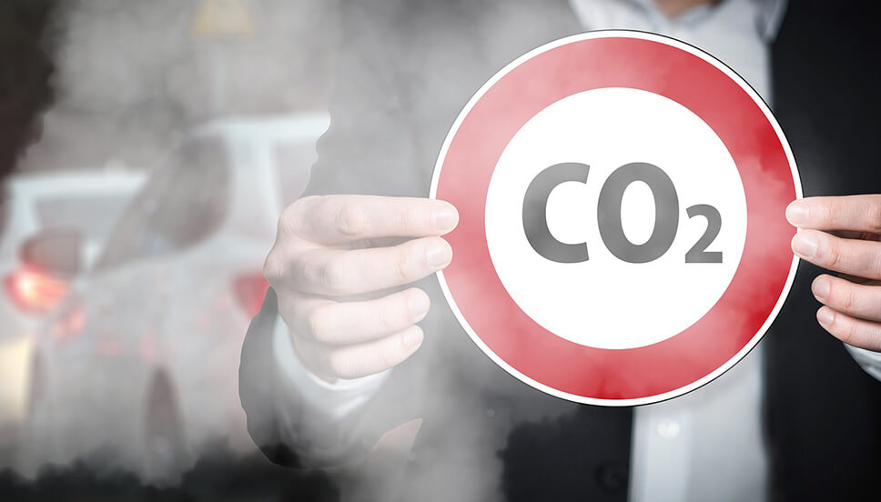 Foto mostrando vários carros e uma fumaça tomando conta da cena, a qual também está presente um homem (em meio à fumaça) segurando uma placa, na qual está escrito CO2