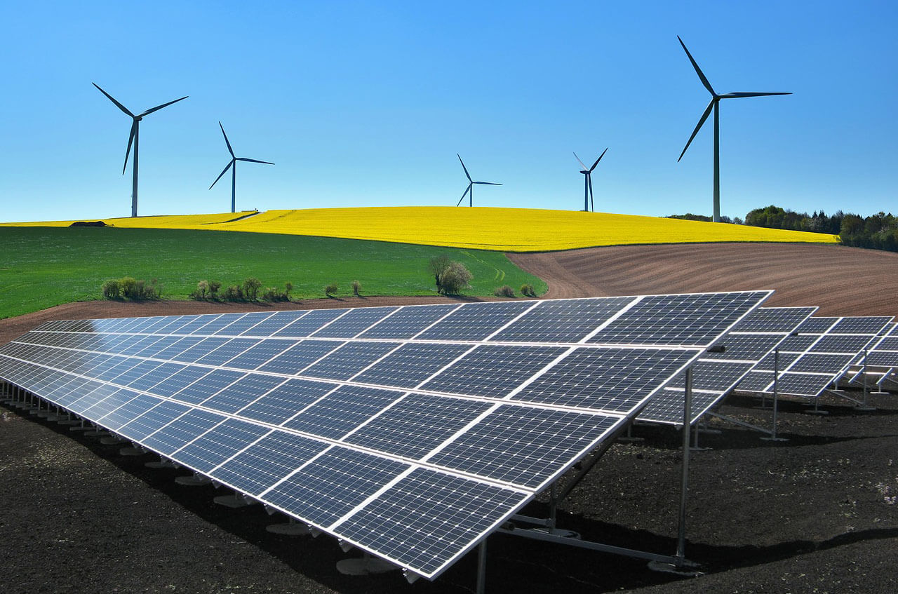 Foto mostrando placas de energia solar (como pano de frente) com cinco geradores de energia eólica ao fundo, posicionadas em uma área gramada, com algumas árvores, sob o céu totalmente azul.
