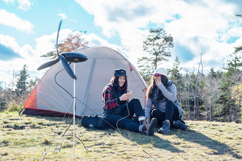 Imagem de duas moças na frente de uma barraca de camping e usando um celular conectado à turbina Shine, ilustrando ser uma turbina eólica portátil que cabe em sua mochila