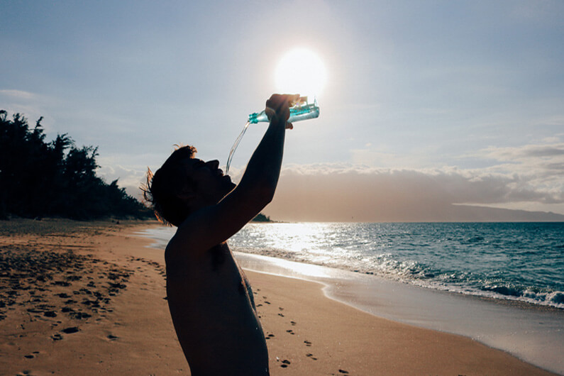 Imagem de um homem na praia, bebendo água em uma garrafa plástica para ilustrar artigo intitulado: você sabe a quantidade de água que precisa beber por dia?