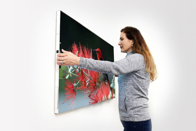 Foto de uma mulher colocando uma TV na parede para ilustrar artigo sobre a primeira TV sem fio e sem controle remoto e que gruda na parede.