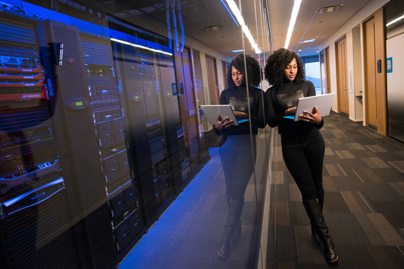 Foto de uma mulher afrodescendente segurando um laptop aberto e posicionada no corredor de uma empresa de tecnologia para ilustrar o artigo sobre como as empresas de tecnologia falham com as mulheres.