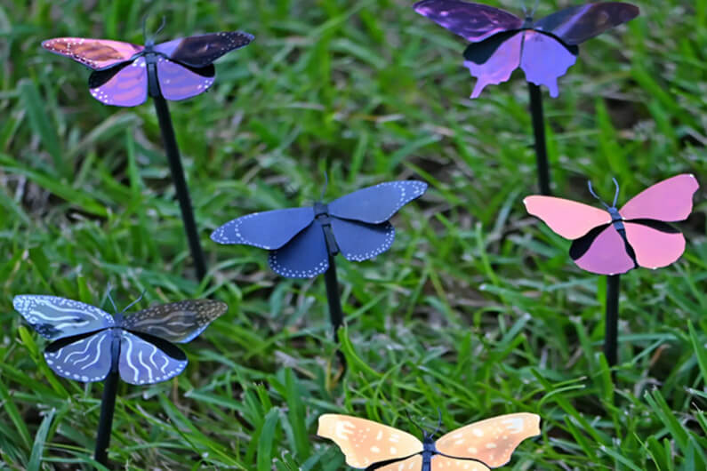 Foto de borboletas metálicas coloridas, fincadas na grama por meio de um ferro, para ilustrar que a tinta plasmática pode tornar a tinta comum coisa do passado.