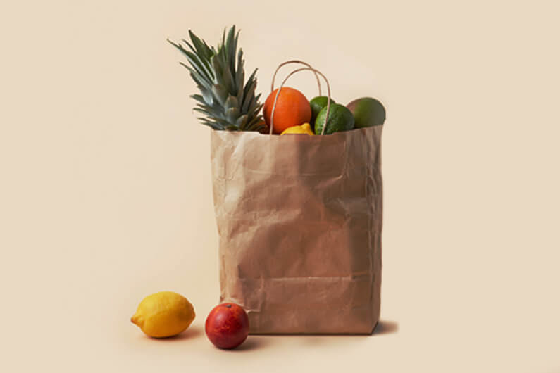 Foto de uma sacola de supermercado, feita de papel, no topo da qual aparecem algumas frutas, e outras do lado de fora da sacola, ilustra artigo que diz que sacola feita de papel forte e barato pode virar biocombustível.