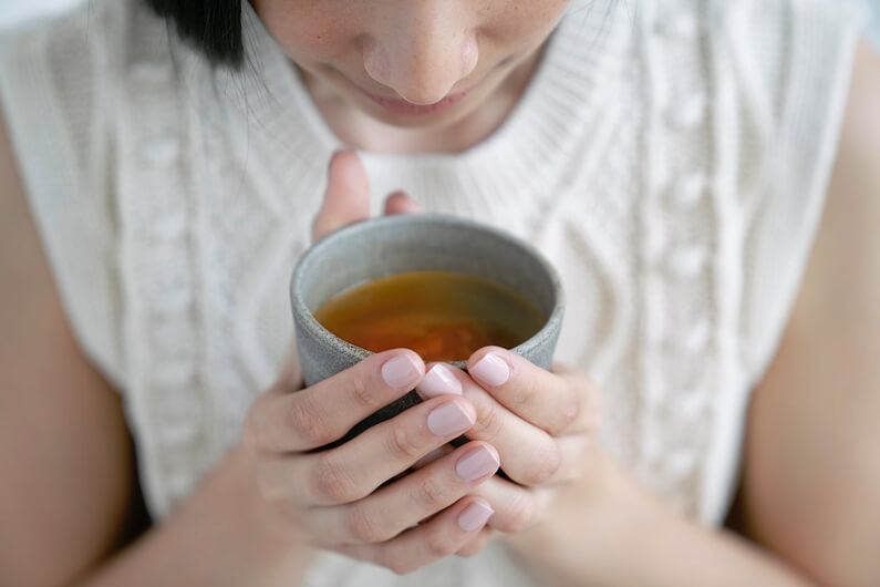 Foto de uma mulher mostrando parcialmente seu rosto olhando para uma xícara de chá que ela segura com as duas mãos para ilustrar que um estudo liga flavonoides à melhora da memória e saúde do coração.