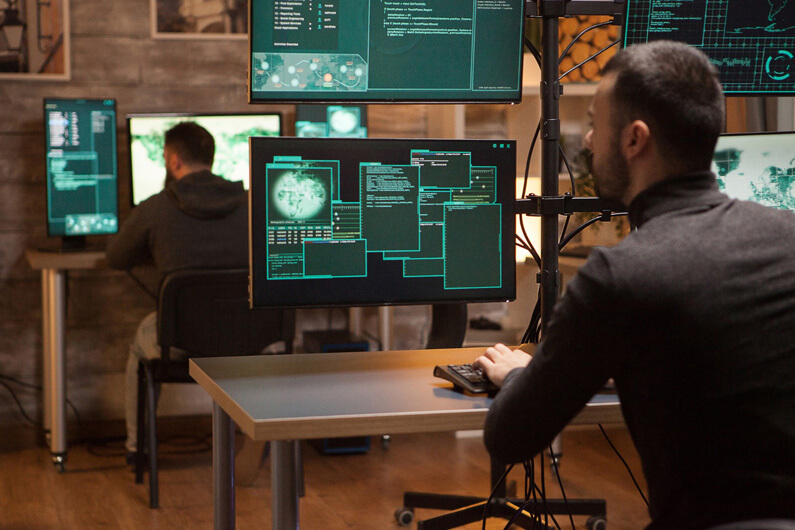 Imagem de uma sala com dois homens olhando para várias telas de monitor para ilustrar o post que indaga sobre se você sabia que seu smartphone é um dispositivo de vigilância.