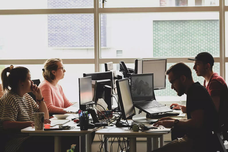 Imagem de uma sala com dois rapazes e duas moças trabalhando na frente de várias telas de computador e laptops para ilustrar o artigo sobre o futuro do emprego na era da sustentabilidade e desglobalização.