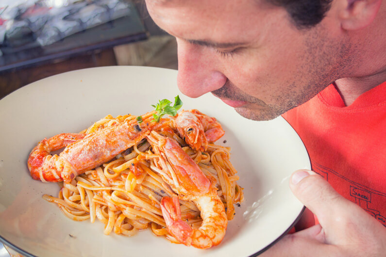 Imagem de um homem reclinando seu rosto sobre um prato de espaguete com camarão para ilustrar o artigo cujo título pergunta: cheirar o alimento é confiável para detectar se ele está estragado?