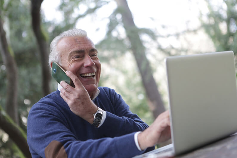Imagem de um senhor ao celular e olhando para a tela de um laptop ilustra que o risco de demência parece diminuir com o uso regular da internet