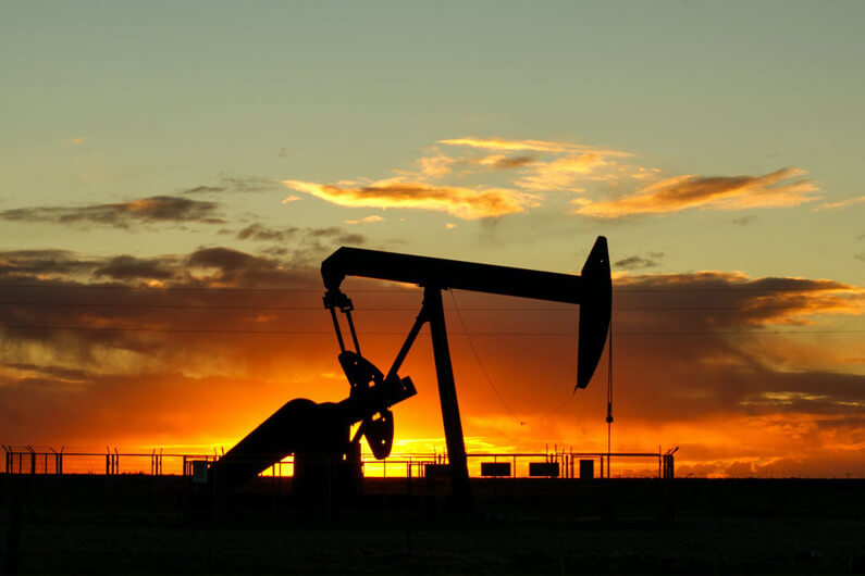 Foto de uma máquina prospectora de petróleo como pano de frente de um campo aberto sob um céu com algumas nuvens e um pôr do sol como pano de fundo ilustra artigo cujo título diz que os combustíveis fósseis atingirão pico até 2030.