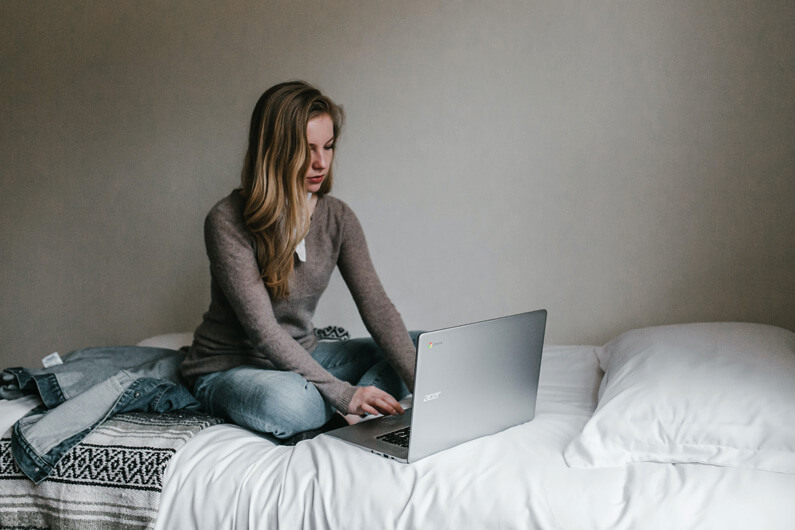 Imagem de uma mulher sentada numa cama e mexendo em laptop aberto para ilustrar artigo cujo título diz que a tecnologia rouba seu tempo de formas que você pode não notar.