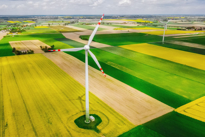 Imagem aérea de uma área aberta e com bastante verde no solo sobre o qual aparecem duas turbinas eólicas ilustram o post cujo título pergunta: como desenvolver as renováveis sem ameaçar a biodiversidade?