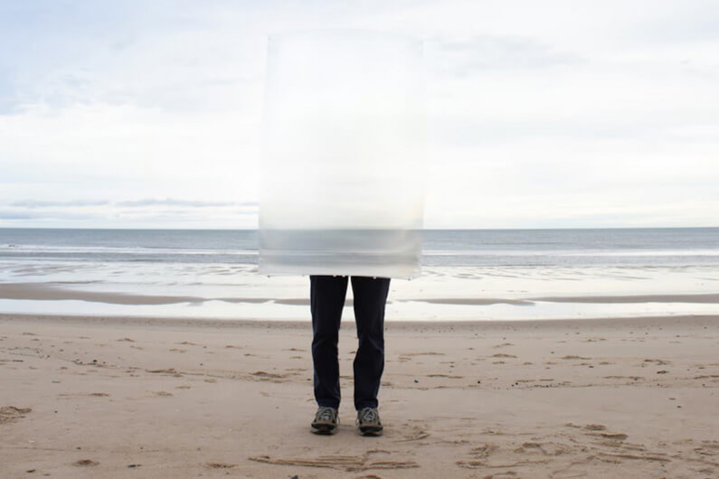 Imagem de um homem na praia segurando um escudo transparente da cintura até a altura acima da cabeça que faz esta parte do corpo ficar invisível com apenas as pernas às vistas serve para ilustrar o post que fala sobre como funciona o escudo que faz as pessoas ficarem 'invisíveis'.