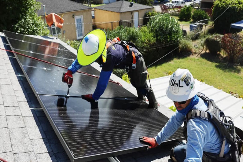 Imagem de dois trabalhadores instalando painéis solares no topo de uma casa ilustra o post cujo título diz que um programa visa tornar energia solar acessível para os mais carentes.